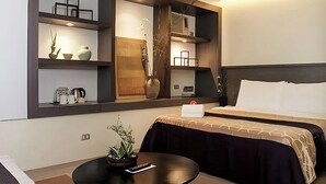 Una habitación de hotel moderna con una cama grande, una mesita de noche con control remoto, una pequeña mesa con una planta y estantes empotrados con objetos decorativos y un florero.