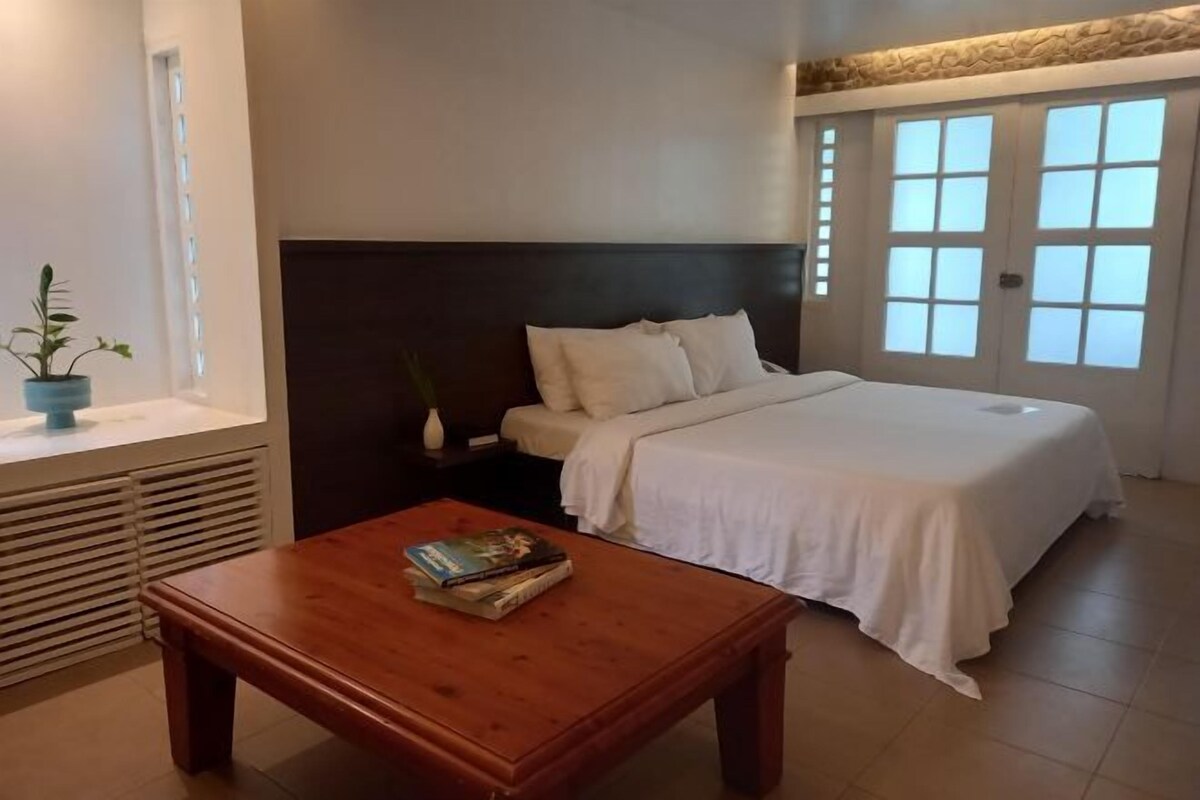 A bedroom with a bed, a bedside table with books, a window with blinds, and a potted plant.