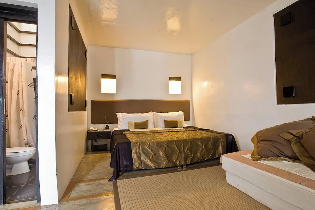A hotel room with a bed, a bedside table, a nightstand, a bathroom with a toilet and shower curtain, and two wall-mounted lights.