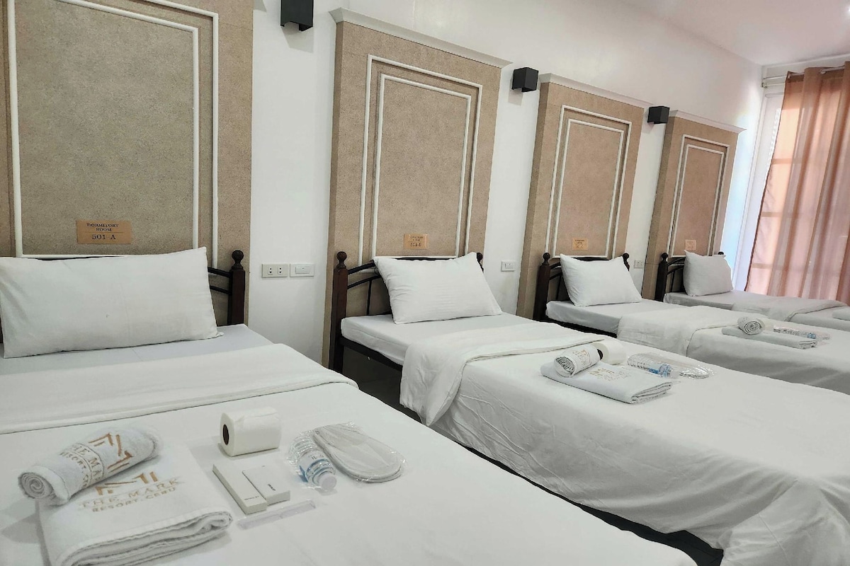 A hotel room with four single beds, each with a white bedspread and pillow, and a small table with toiletries.