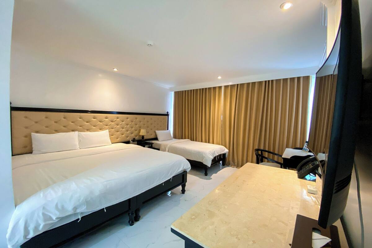 A hotel room with a large bed, two smaller beds, a desk, and a television.