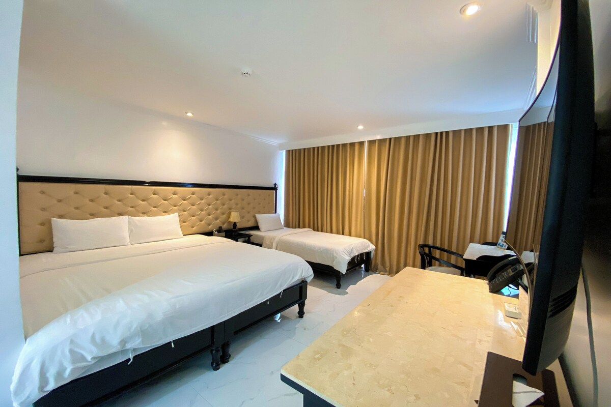 A hotel room with a large bed, two smaller beds, a desk, and a television.