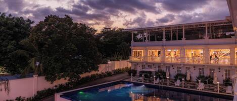 A hotel with a pool, a second-story balcony with outdoor seating, and a large tree in the background.