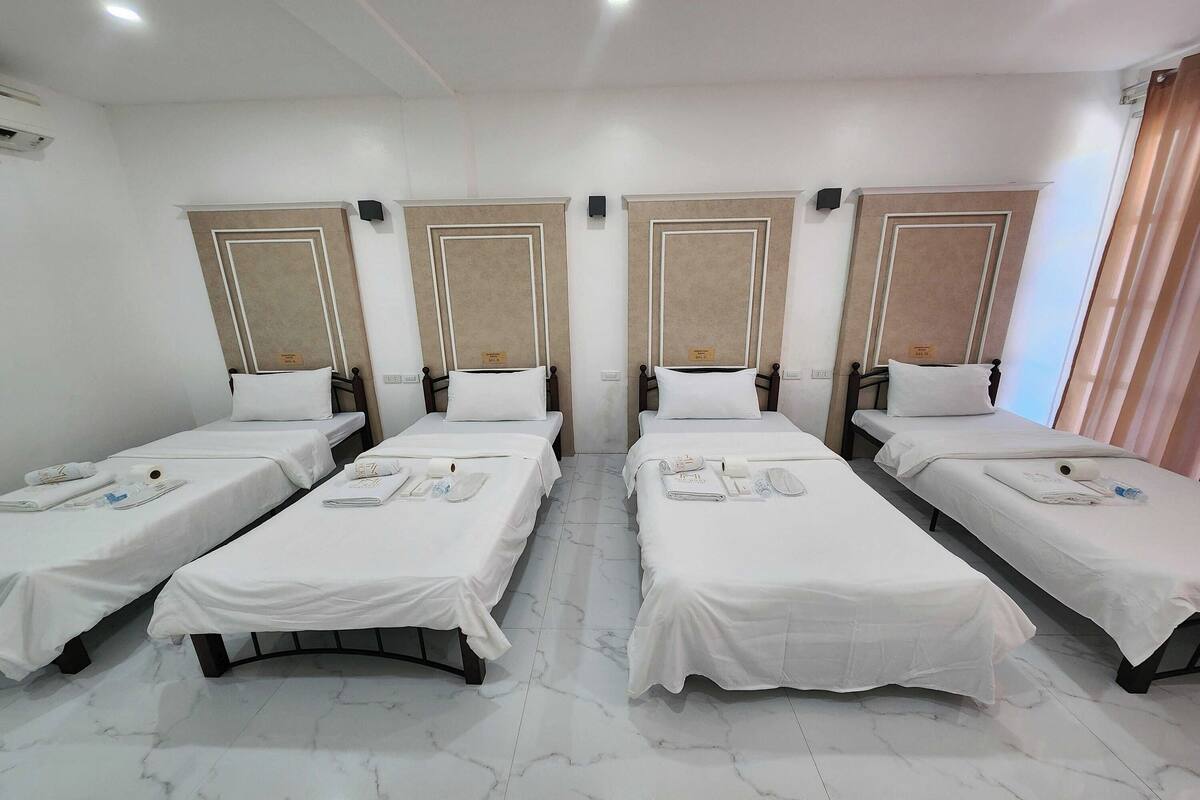 A hotel room with four single beds, each with a white bedspread and a pillow. The room has a marble floor and beige headboards with decorative trim.