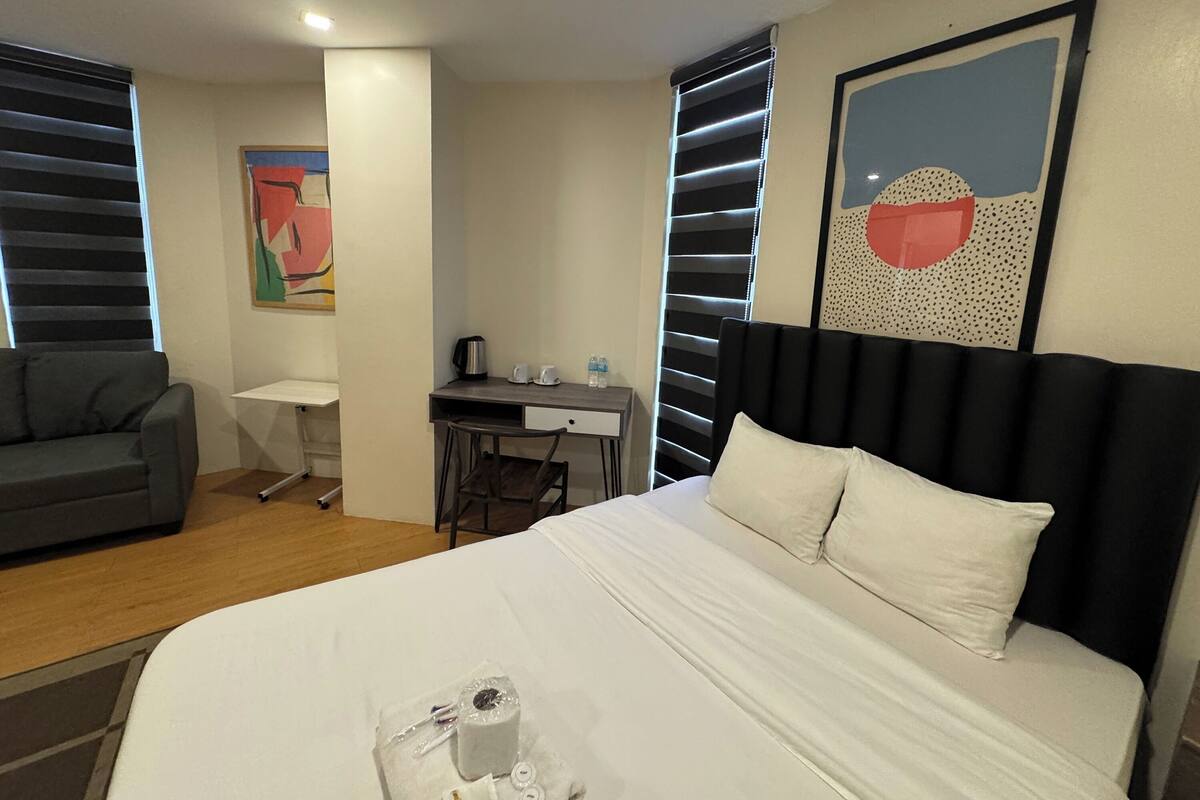 A hotel room with a bed, a desk, a chair, a sofa, and artwork on the walls.