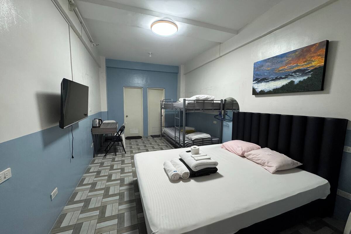 A hotel room with a bunk bed, a single bed, a flat-screen TV, a painting, and a tiled floor.