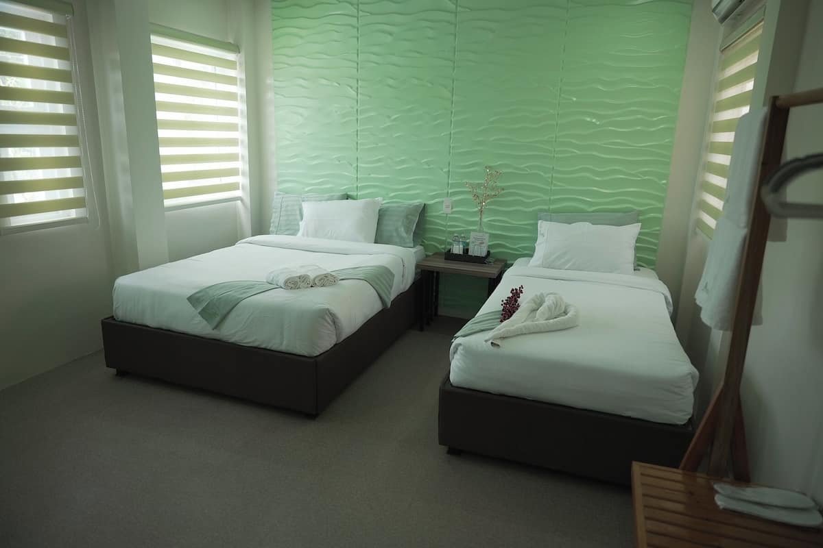 A modern hotel room with two beds, a green textured wall, and a small bedside table with a vase.