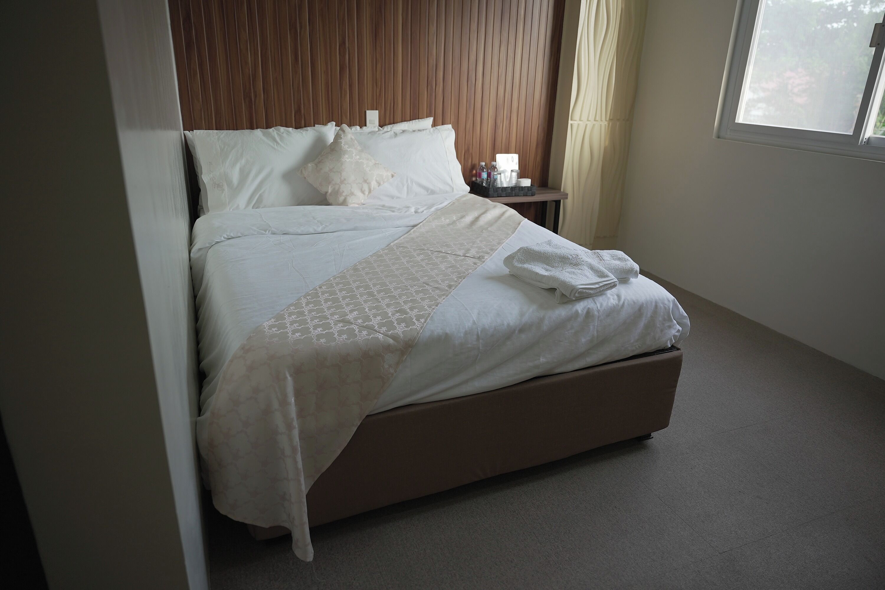 Una cama bien hecha con una colcha estampada y almohadas, una mesita de noche con artículos de aseo y una ventana con cortinas.