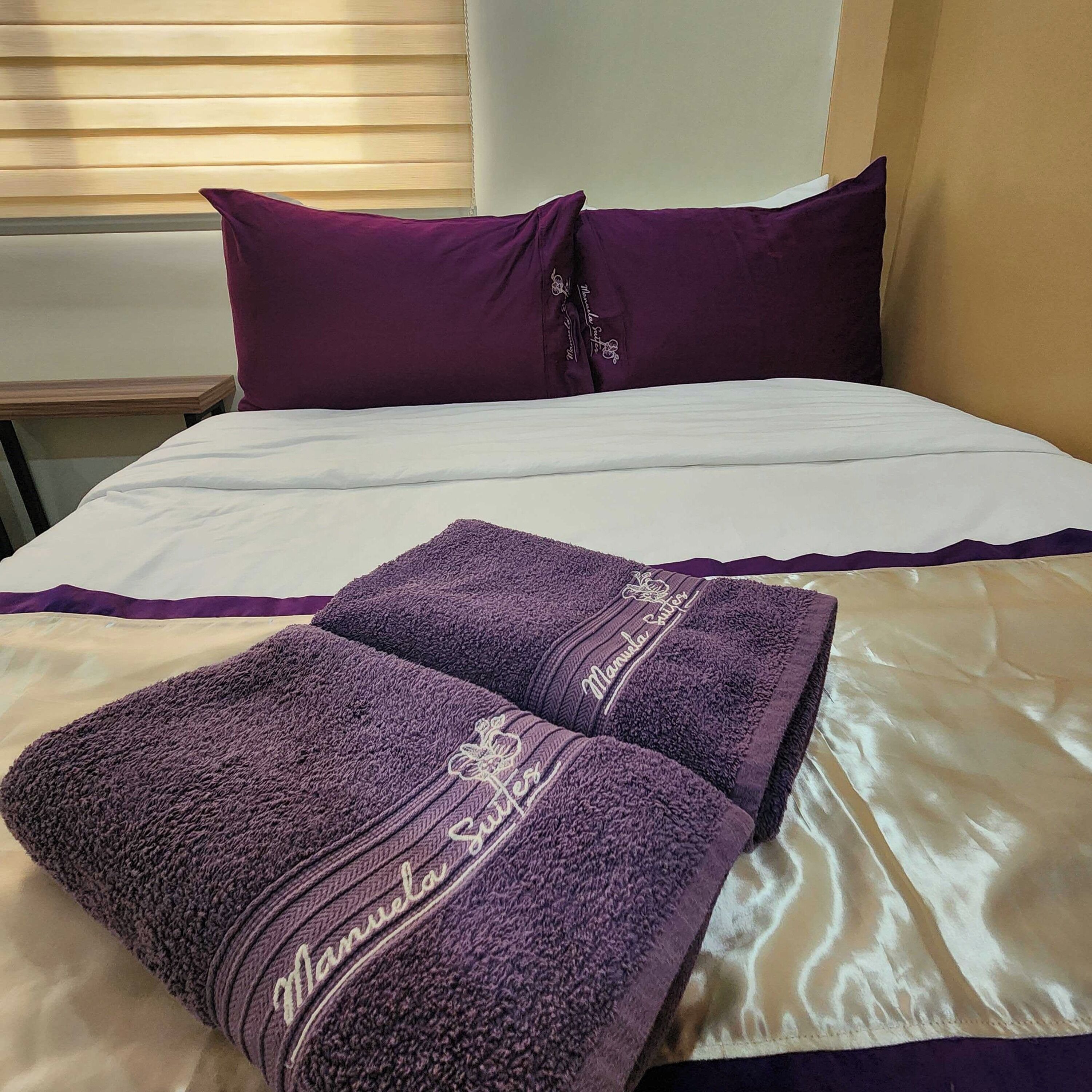Ein ordentlich bezogenes Bett mit weißer und violetter Bettwäsche, zwei violette Handtücher mit eingesticktem Text, und ein Fenster mit Jalousien im Hintergrund.