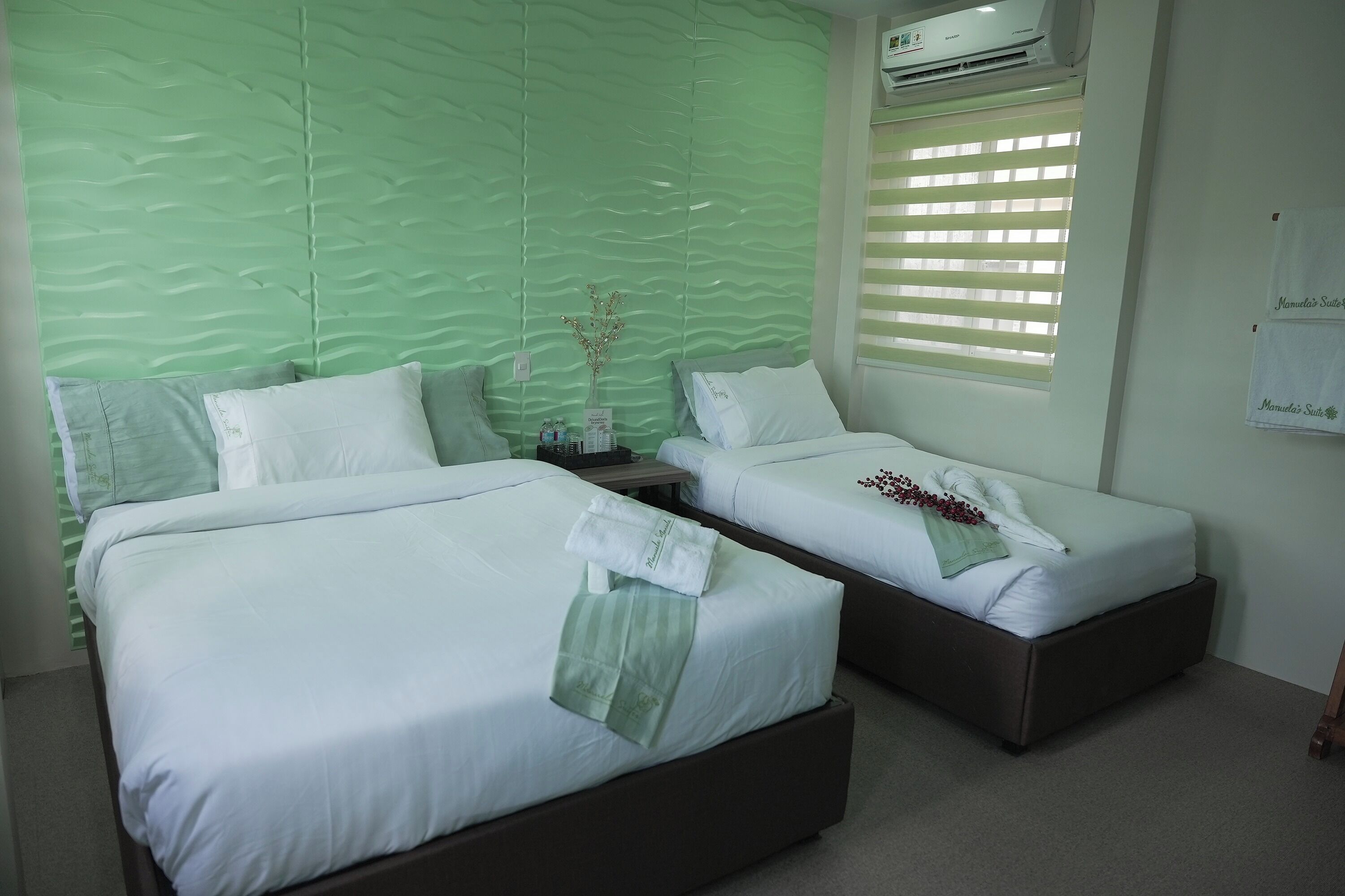 Habitación de hotel con dos camas, una pared verde con textura, una ventana con persianas y un aire acondicionado de pared.