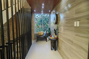 A modern interior with a wooden floor, a black metal partition, a wooden wall with a geometric pattern, a wooden ceiling, recessed lighting, a window with a view of greenery, a chair with cushions, and a small table with a plant.