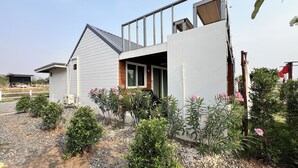 A modern two-story house with a balcony, surrounded by various shrubs and small plants.