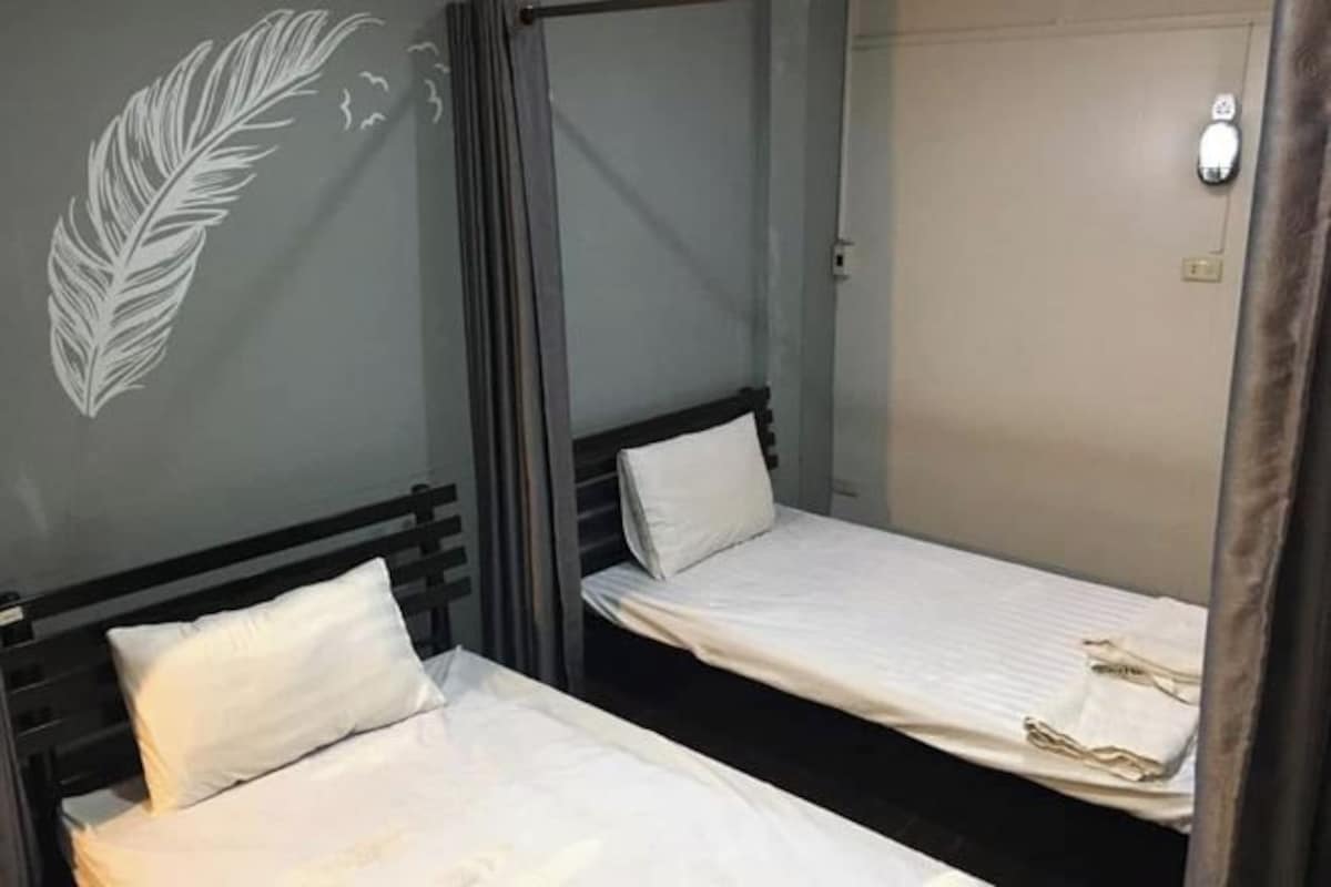 Une chambre avec deux lits simples, un mur gris orné d’un motif blanc en forme de plume et une porte avec un interrupteur.