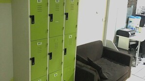 A room with green lockers labeled B1 to B8, a brown sofa, and a desk with a computer and printer. - Ali Hostel (Bangkok)