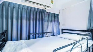 A single bed with a metal frame, a white mattress, and a white pillow. There is a window with blue curtains, and an air conditioning unit mounted on the wall above the bed. - Ali Hostel (Bangkok)