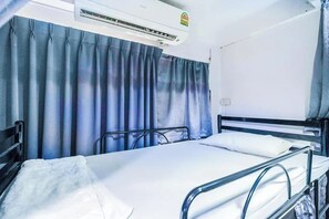 A single bed with a metal frame, a white mattress, and a white pillow. There is a window with blue curtains, and an air conditioning unit mounted on the wall above the bed. - Ali Hostel (Bangkok)
