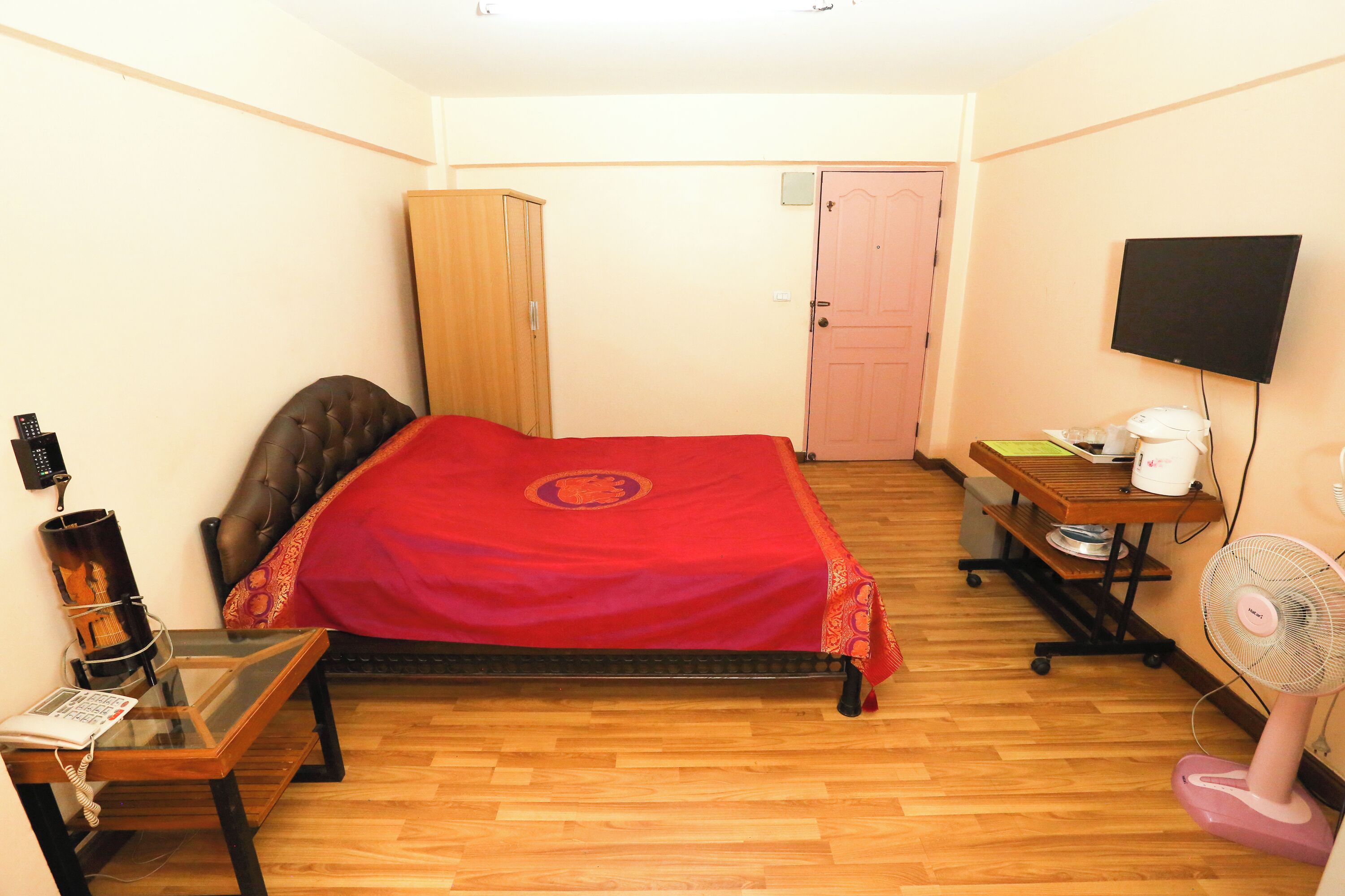 A small, well-lit room with a single bed, a wooden wardrobe, a desk with a chair, a television, and a standing fan.