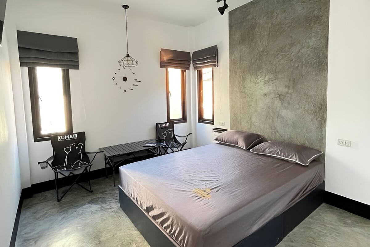 A bedroom with a bed, two windows with curtains, a desk with a chair, and a hanging light fixture.