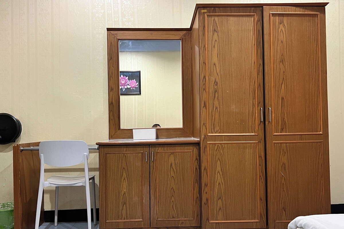 A room with a wooden wardrobe, a desk with a chair, and a bed.