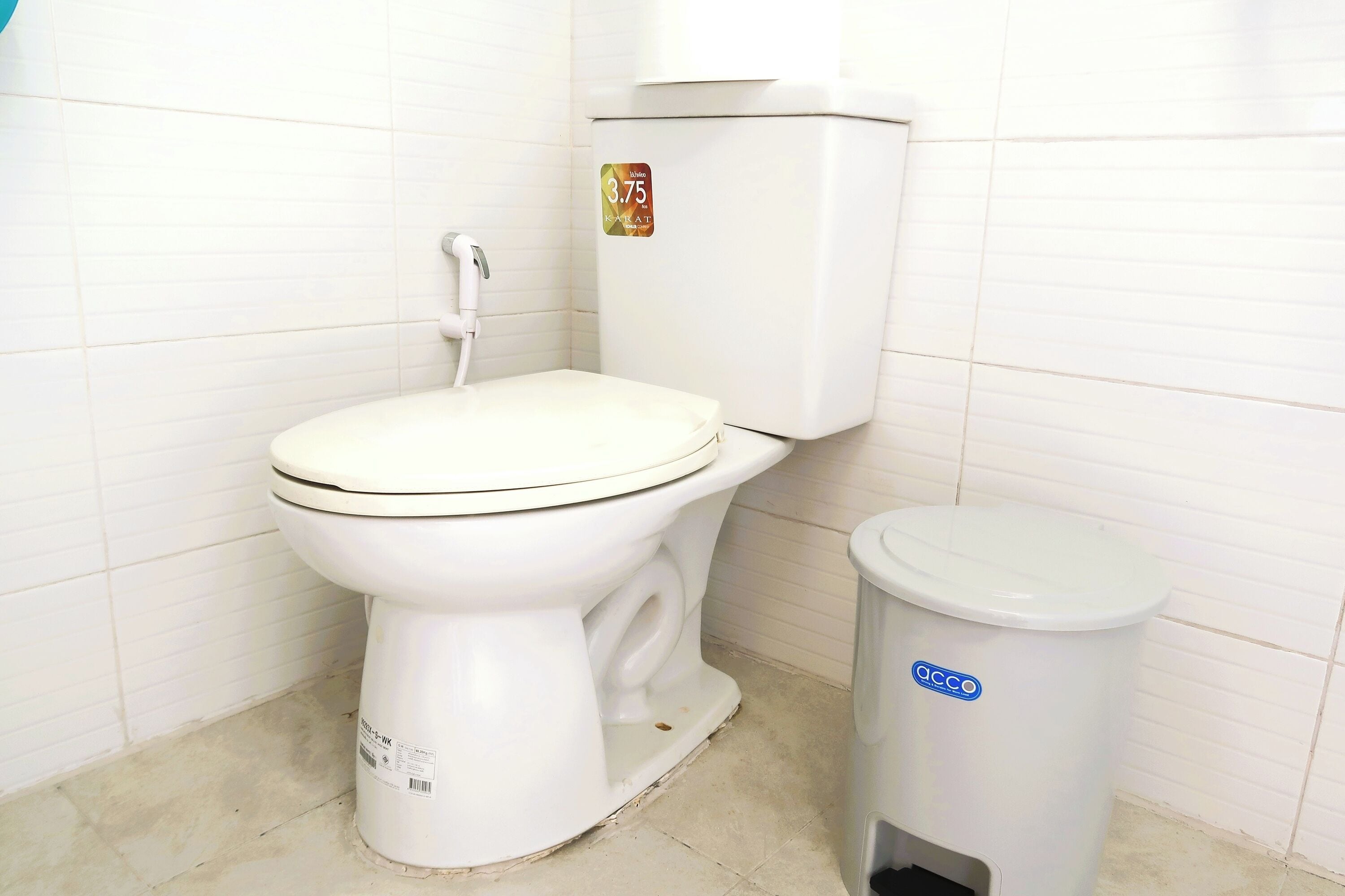 Un inodoro con asiento y tanque blancos, un cubo de basura blanco con logotipo azul y paredes con azulejos.