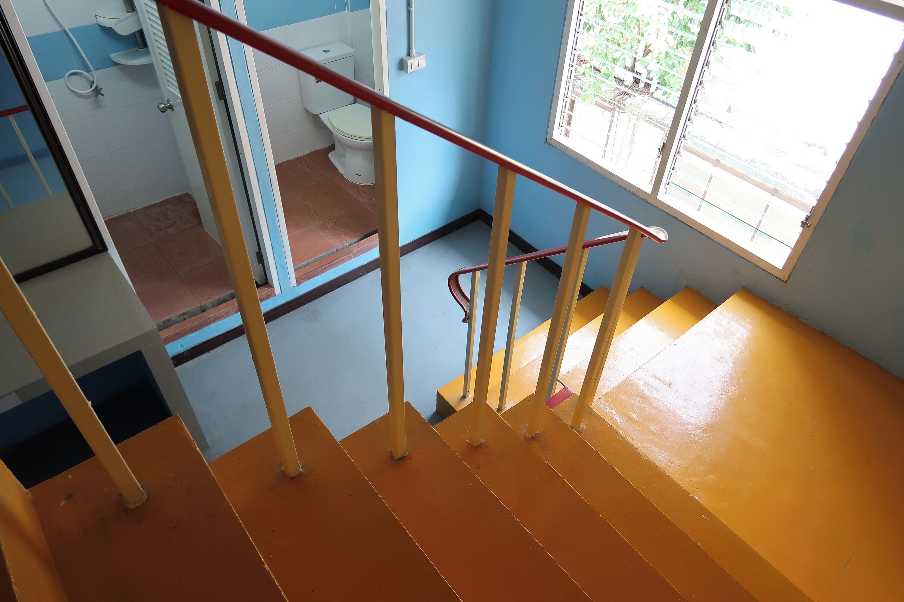 Una escalera con peldaños amarillos y barandilla roja que conduce a una habitación con inodoro y ventana.