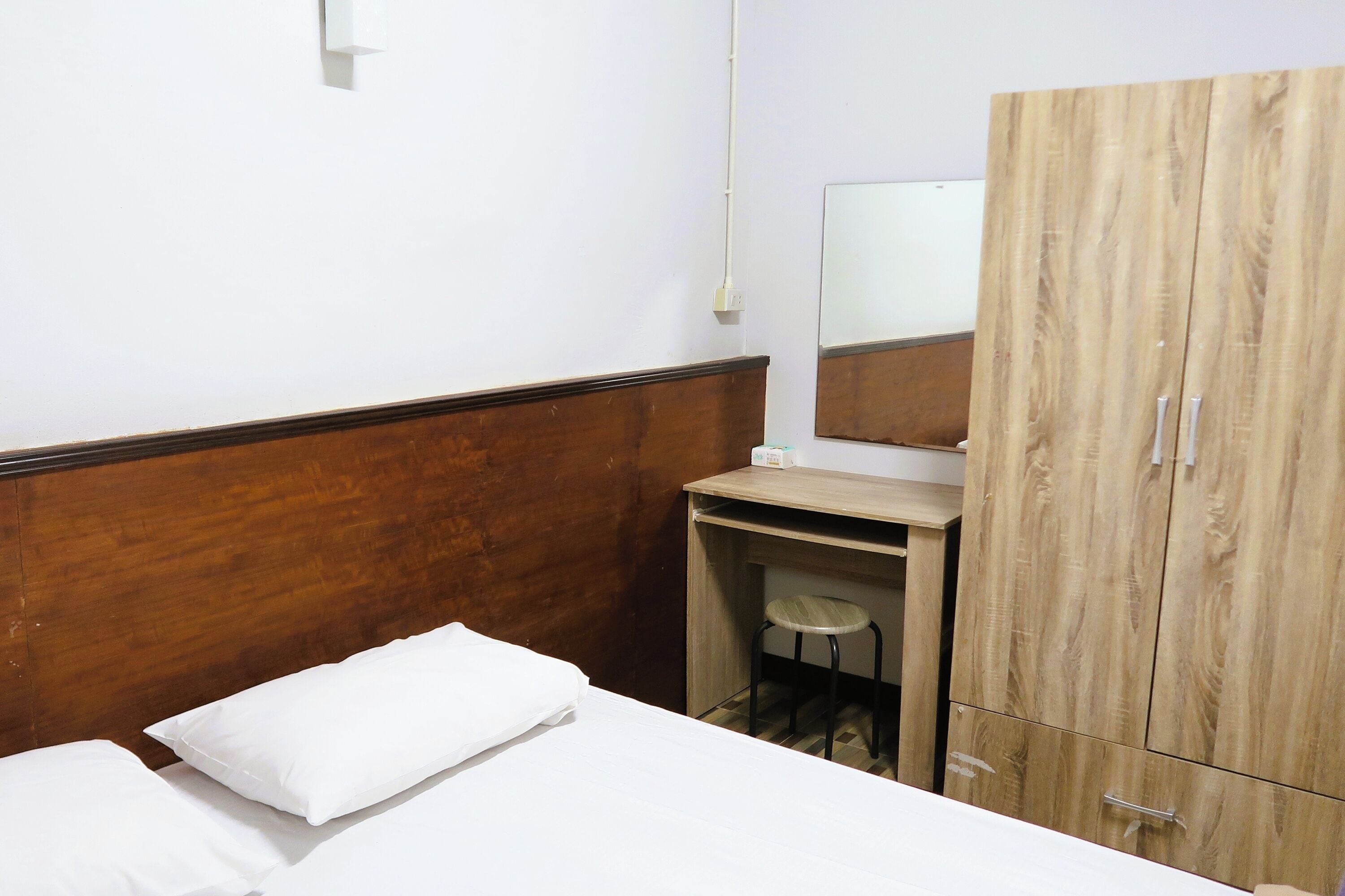 A hotel room with a wooden headboard, a bed with white linens, a small desk with a stool, and a wardrobe with a mirror.