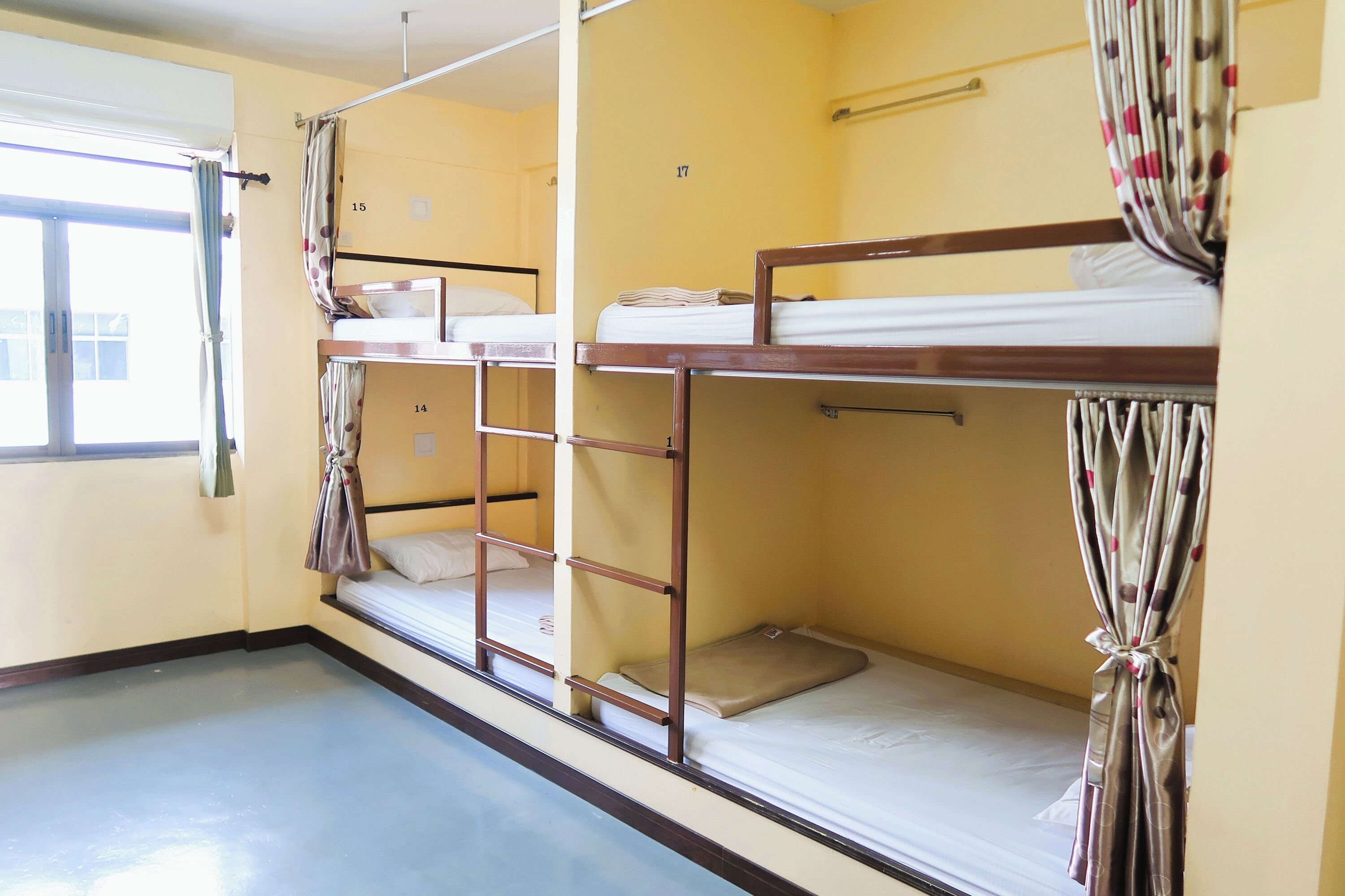 A dormitory room with bunk beds, a window with curtains, and a door.