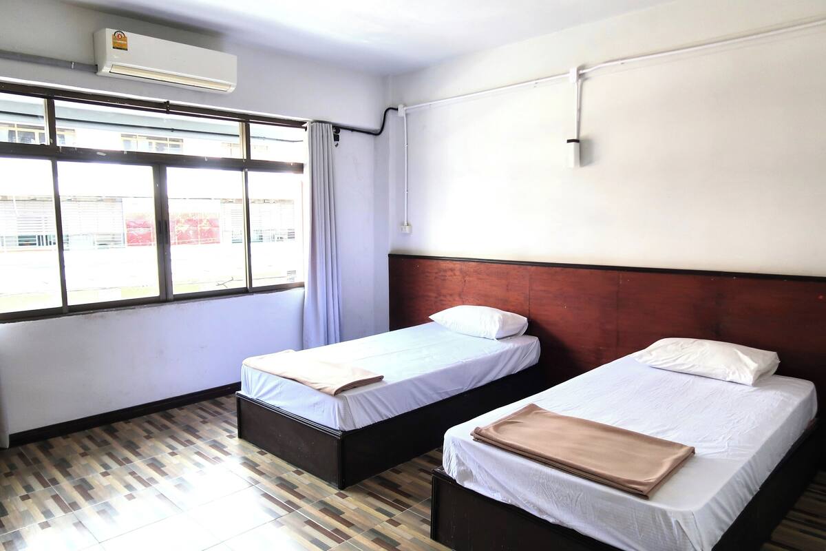 Een kamer met twee bedden, een raam met gordijnen, een wandairconditioner en een betegelde vloer.