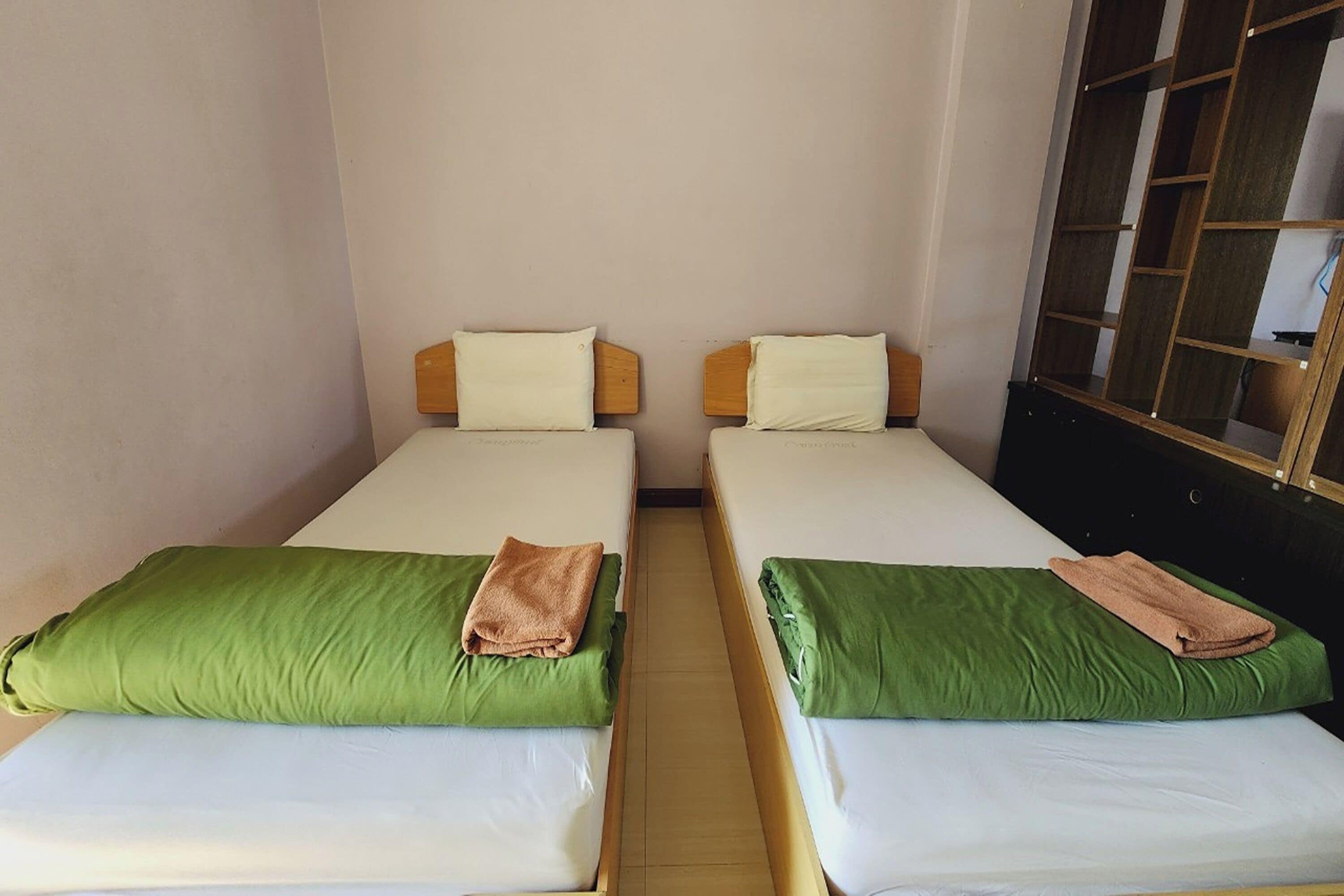 Two single beds with green and white bedding, wooden headboards, and a small wooden shelf unit to the right.