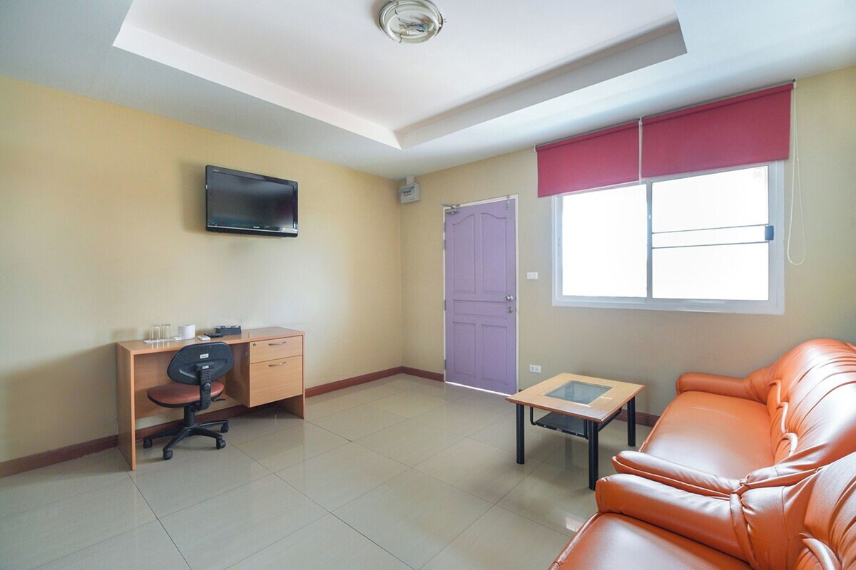A room with a desk, office chair, sofa, TV, and a window with a red blind.
