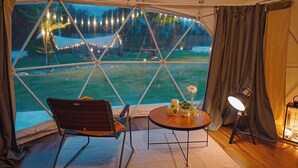 A tented room with a view of a lake, a round table, two chairs, a lamp, and a flower arrangement. - Nanine Glamping By Zuzu (Cha-am)