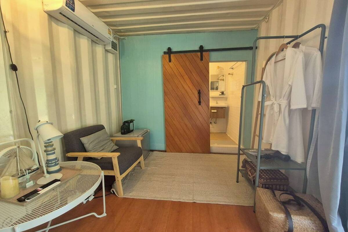 A compact room with a sofa, a small table with a lamp, a sliding door leading to a bathroom, and a rack with towels.
