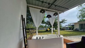 A modern outdoor bathtub with a white exterior, surrounded by a covered patio area with white curtains and hanging pendant lights. - Nanine Glamping By Zuzu (Cha-am)