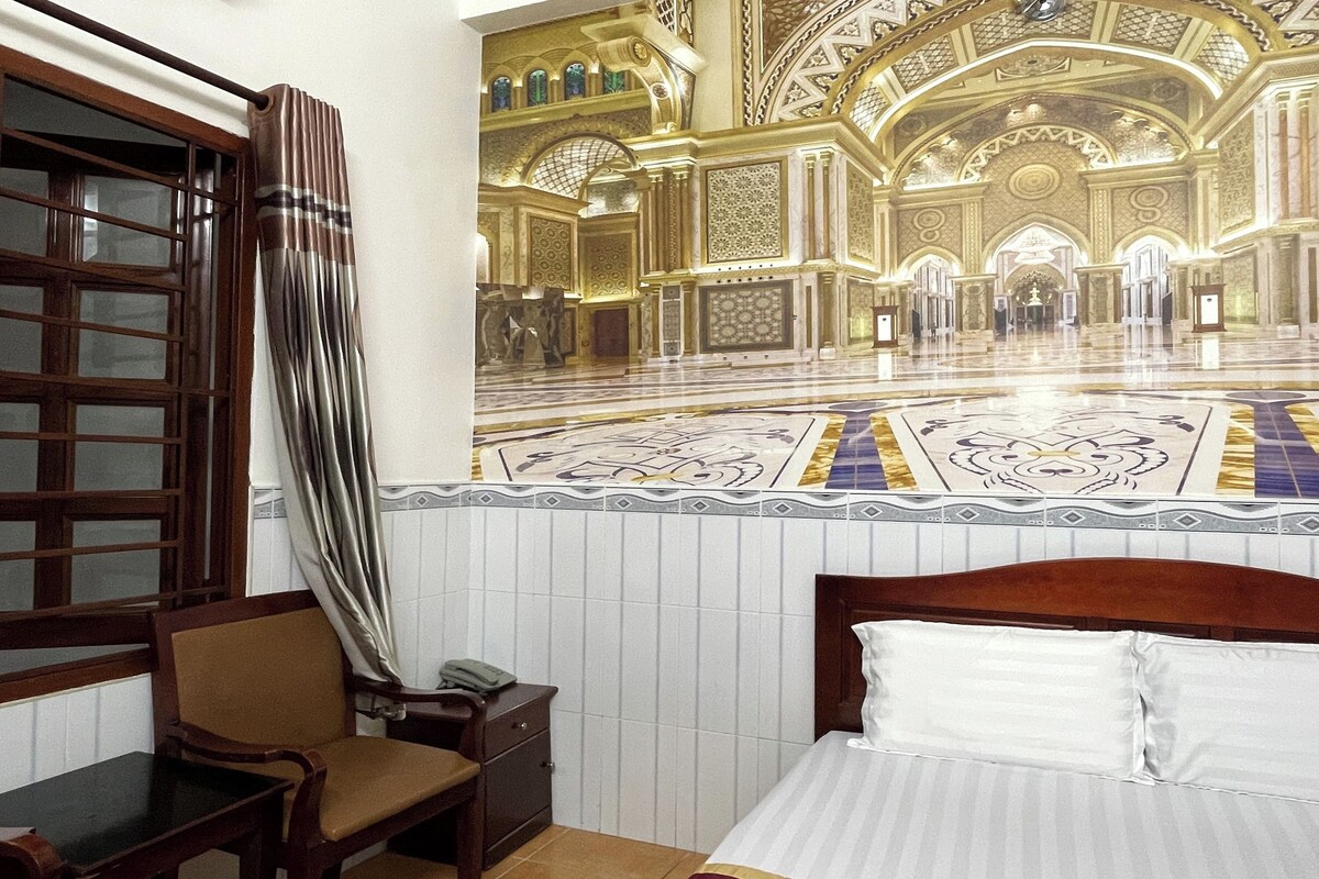 Ein Hotelzimmer mit einem großen Bett, einem Holfnachtkästchen, einem Stuhl und einem Vorhangfenster.