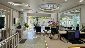 Un moderno vestíbulo de hotel con un mostrador de recepción, un televisor y una computadora portátil sobre la mesa. Se observan plantas en macetas y un ventanal grande al fondo.