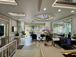 Um lobby de hotel moderno com uma mesa de recepção, uma televisão e um laptop sobre a mesa. Há plantas em vasos e uma grande janela ao fundo.
