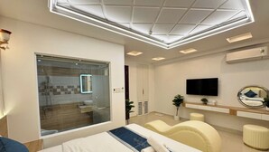 Habitación de hotel moderna con una cama grande, televisión, zona de estar y baño visible a través de un panel de vidrio.