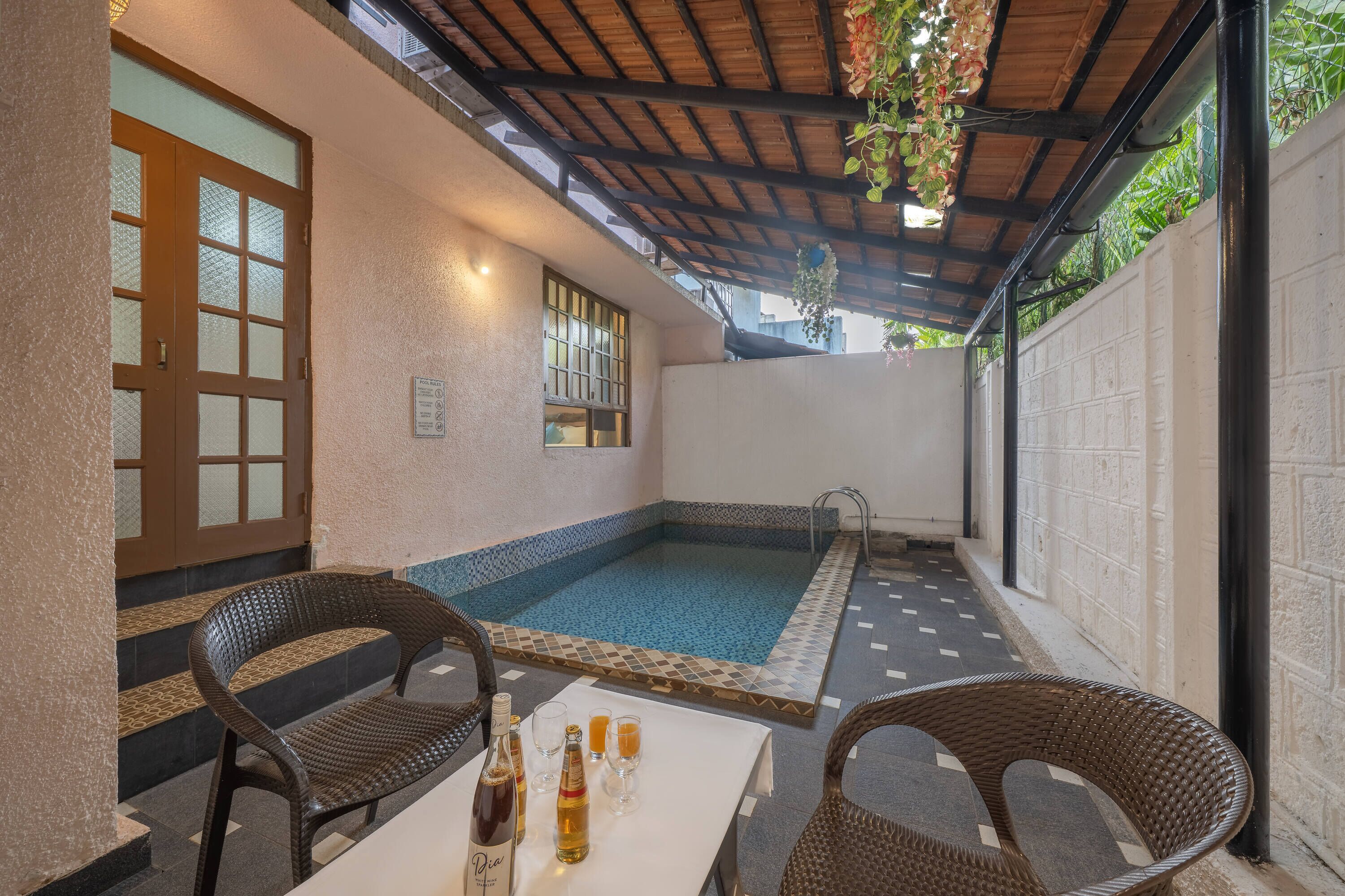 A covered patio area with a pool, wicker chairs, and a table with bottles and glasses.