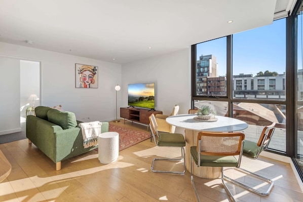 A modern living room with a green sofa, a round dining table, and a large window offering a city view.