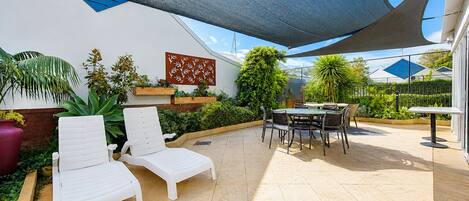 A covered patio area with a table and chairs, a white lounge chair, and various plants.