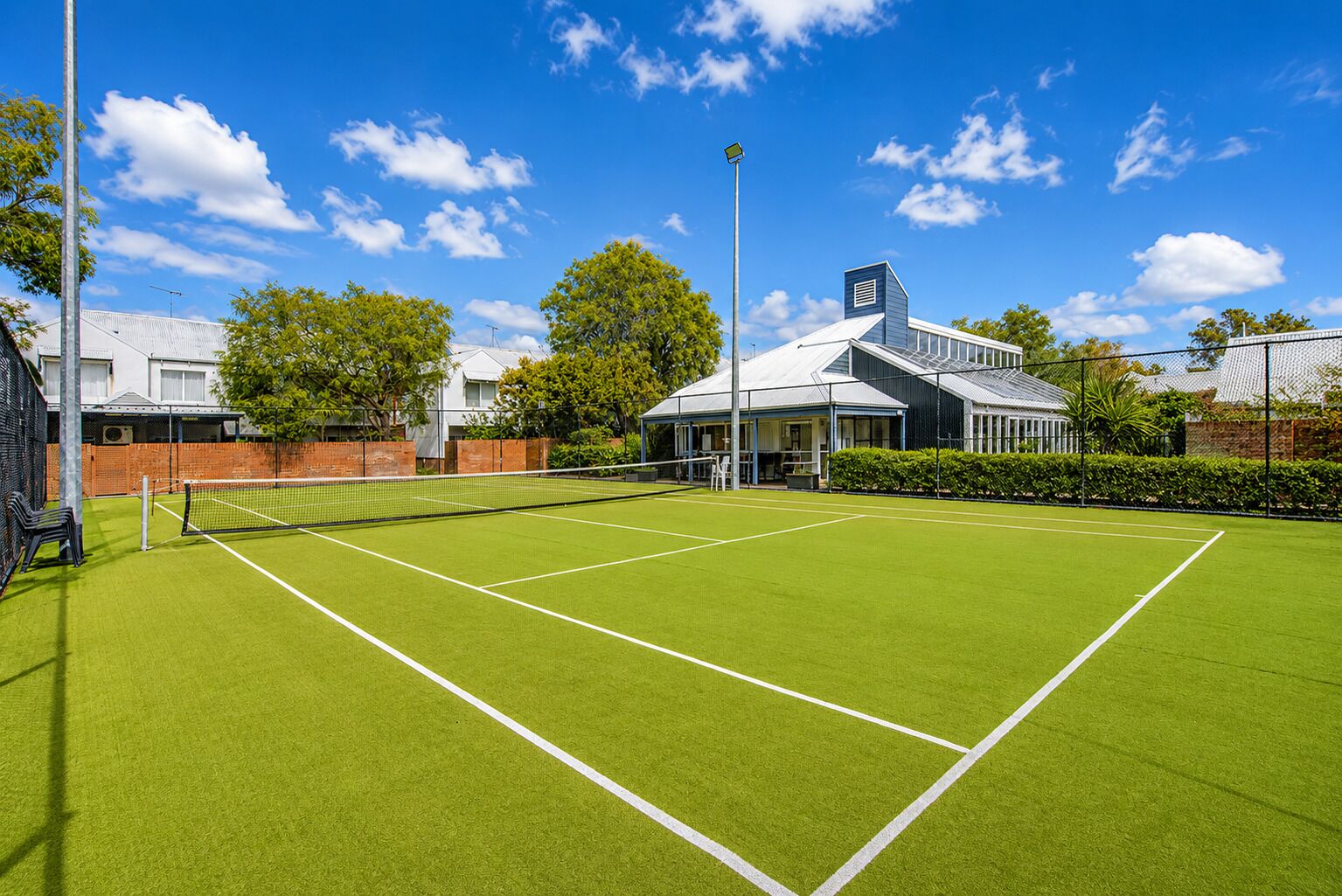 A tennis court with a net, surrounded by a fence, and adjacent to a building with a covered seating area.