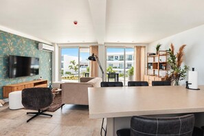 A modern living room with a flat-screen TV, a sofa, a dining table with chairs, and a large window with a view of palm trees and buildings.