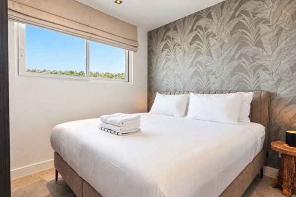 A neatly made bed with white linens and pillows, a window with a view, and a patterned wallpaper on the wall.