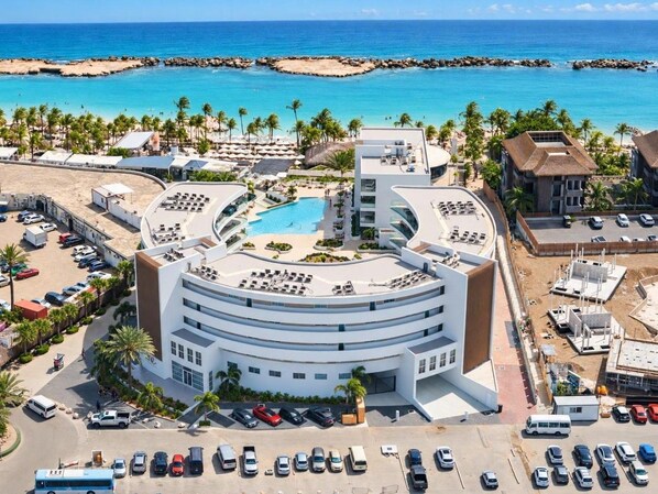 A modern hotel with a circular pool, a beach view, and a parking lot.