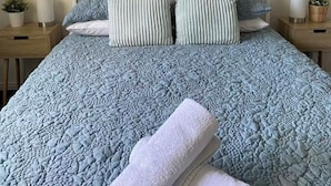 A neatly made bed with a blue quilt, two pillows, and rolled towels on a white metal headboard.
