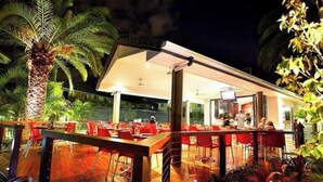 An outdoor dining area with red chairs and tables, surrounded by palm trees and plants.