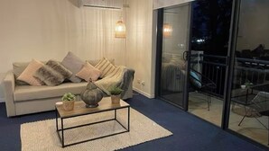 A living room with a sofa, coffee table, and potted plants. There is a sliding glass door leading to an outdoor area with chairs.