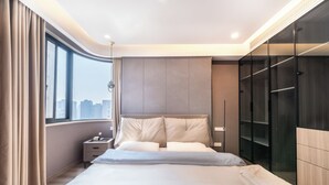 A modern hotel room with a large bed, a nightstand, a wardrobe, and a view of the city through large windows.
