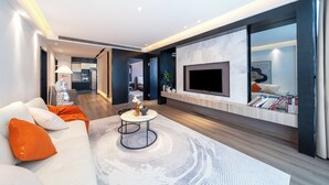 A modern living room with a white sofa, orange pillows, a round coffee table, and a large flat-screen TV mounted on a marble wall.