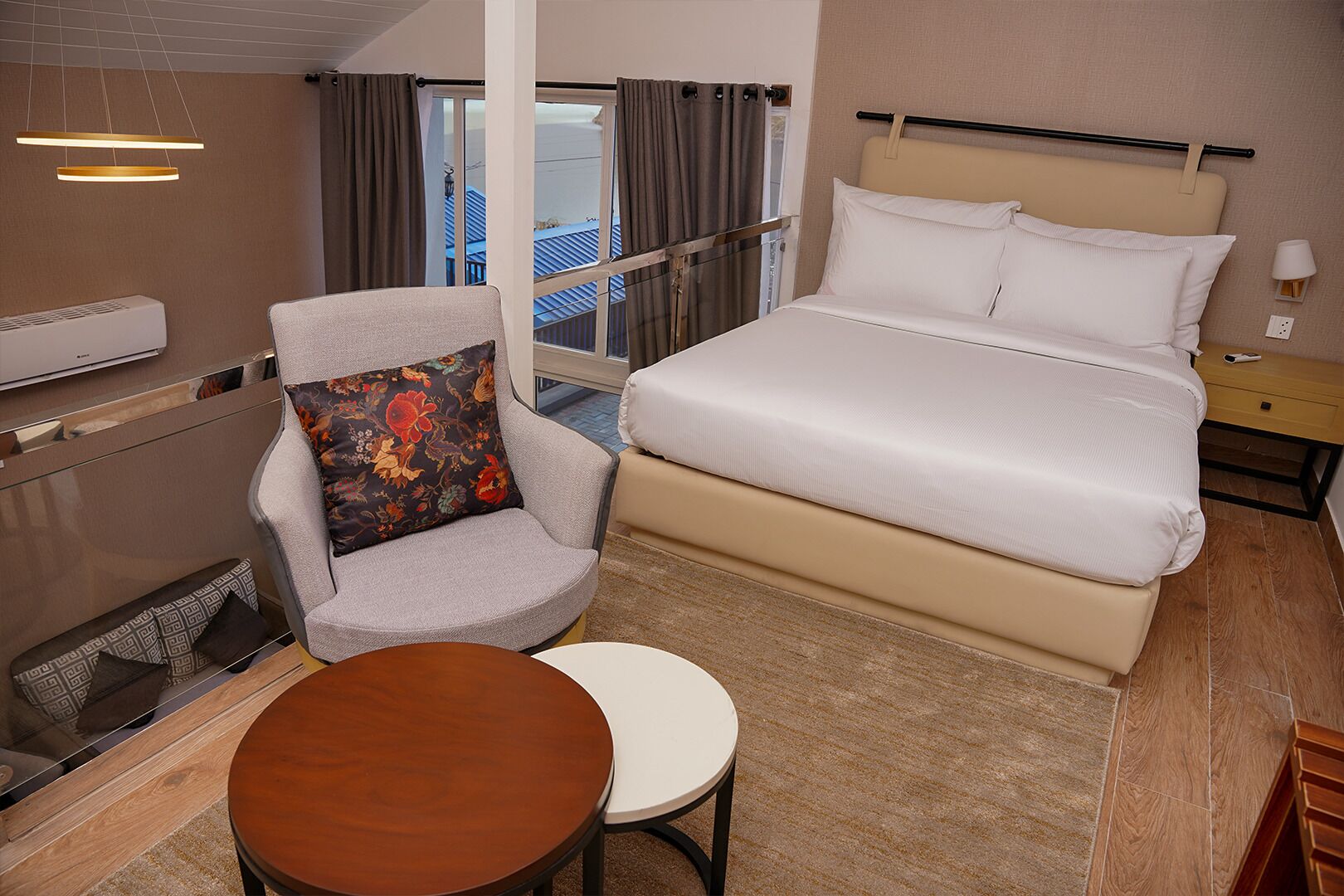 Une chambre d’hôtel avec un lit, une chaise agrémentée d’un coussin à motifs floraux, une petite table ronde et une fenêtre avec des rideaux.